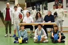 Jürgen Poerschke (am Tisch, links) und Michael Walter (am Tisch, rechts) besiegelten mit ihrer Unterschrift und im Beisein von Projektkoordinatorin Claudia Geiger (am Tisch, Mitte), Steve Bopp (links) und der Nachwuchs-Fechter die weitere Kooperation. Zwei Männer und eine Frau sitzen an einem Tisch, umgeben von Kindern und Jugendlichen mit Fechtausrüstung.
